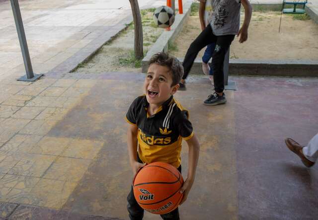 Ein Kind hält einen Basketball in einem überdachten Außenbereich, während im Hintergrund weitere Personen zu sehen sind.
